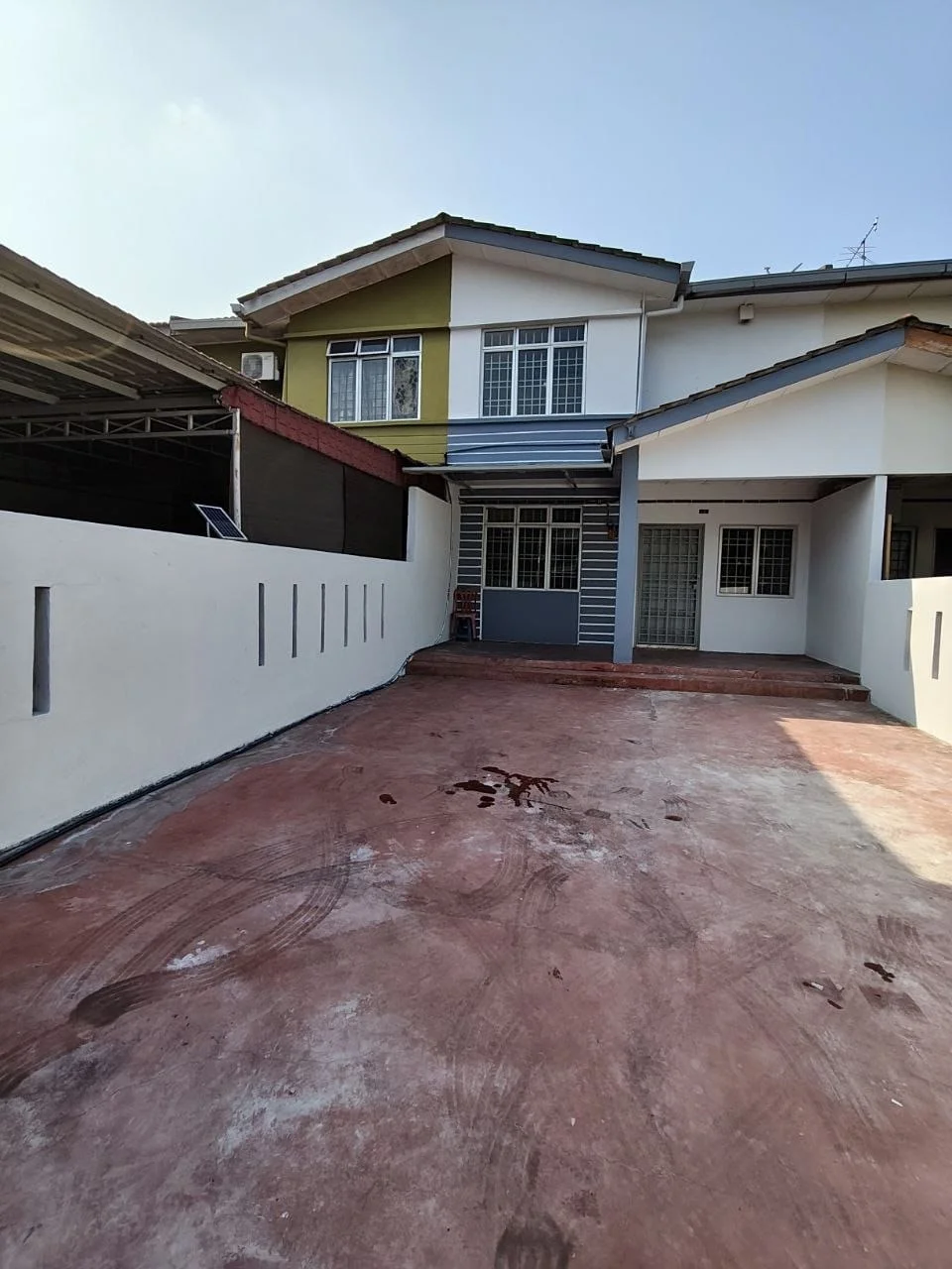 Exterior type: linked terrace house; features: two-storey building, painted facade in yellow and grey, concrete driveway, manicured lawn area, car porch, windows with grilles, main entrance door, exposed brick wall, roof overhang, surrounded by low fencing; style: modern.